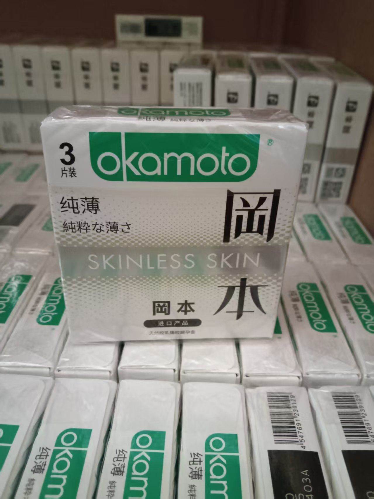 Real stock display: a complete green-logo Okamoto Pure Thin 3-piece carton stands among matching cartons; foreground stock barcode 4547691239129 is readable.
